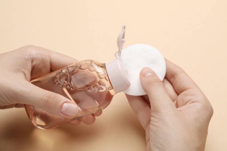 Woman pouring micellar water from bottle onto cotton pad against beige background
