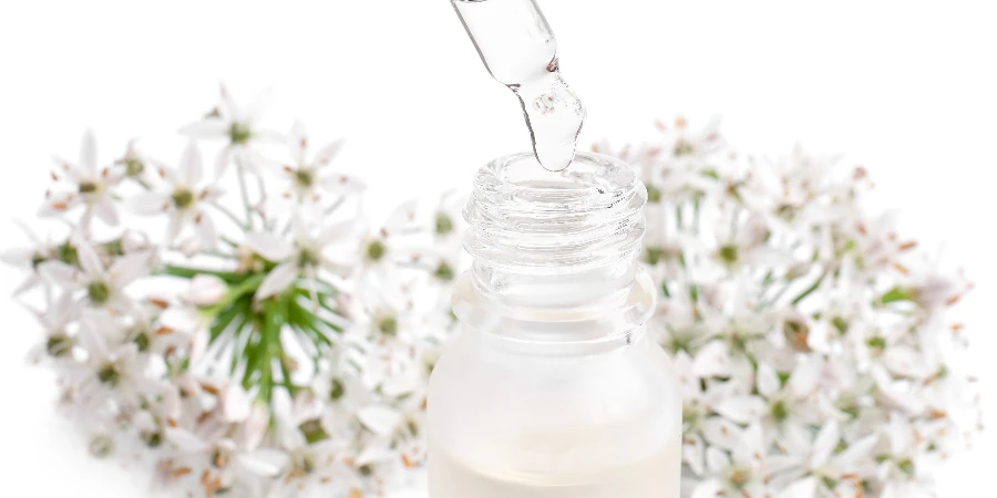 Dripping essential serum from pipette into bottle near garlic chives flowers on white background