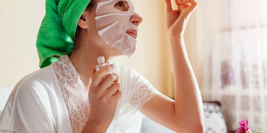 Woman applying facial hyaluronic acid serum wearing sheet mask and towel after shower