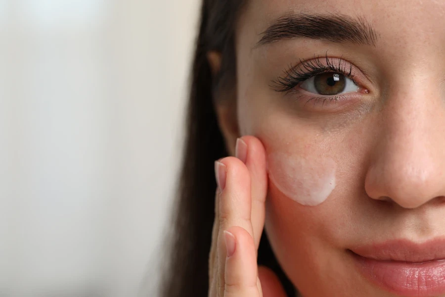 Young woman with dry skin applying cream onto her face on blurred background
