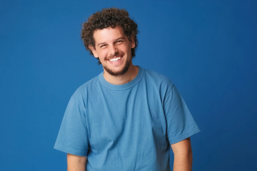 Happy guy with curly hair dressed in blue t-shirt laughing looking at camera isolated on blue background in studio
