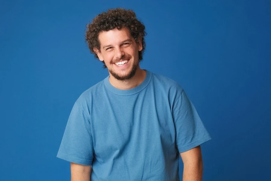 Happy guy with curly hair dressed in blue t-shirt laughing looking at camera isolated on blue background in studio