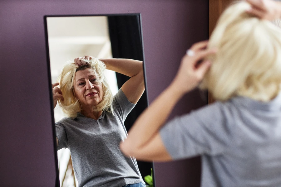 Portrait of mature bald woman putting on wig looking in mirror while getting ready in morning