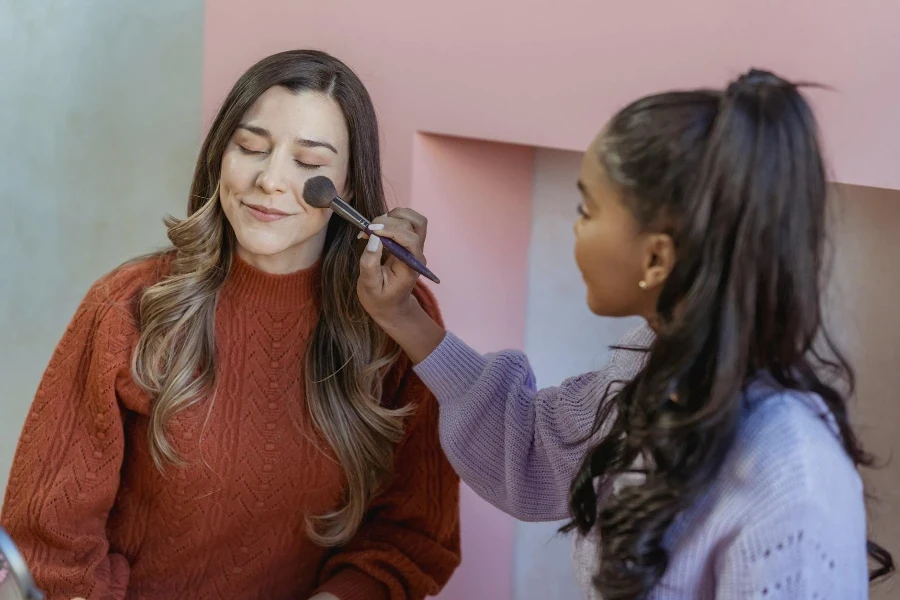 Young ethnic lady applying foundation on face of friend with brush