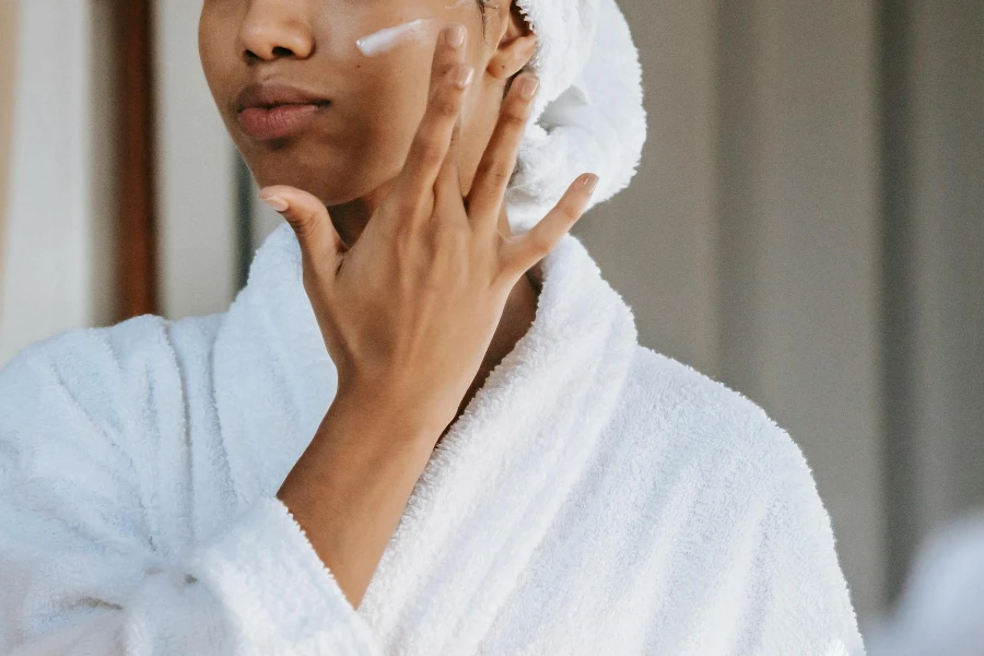 Crop anonymous female in white bathrobe with towel on head applying facial moisturizing cream