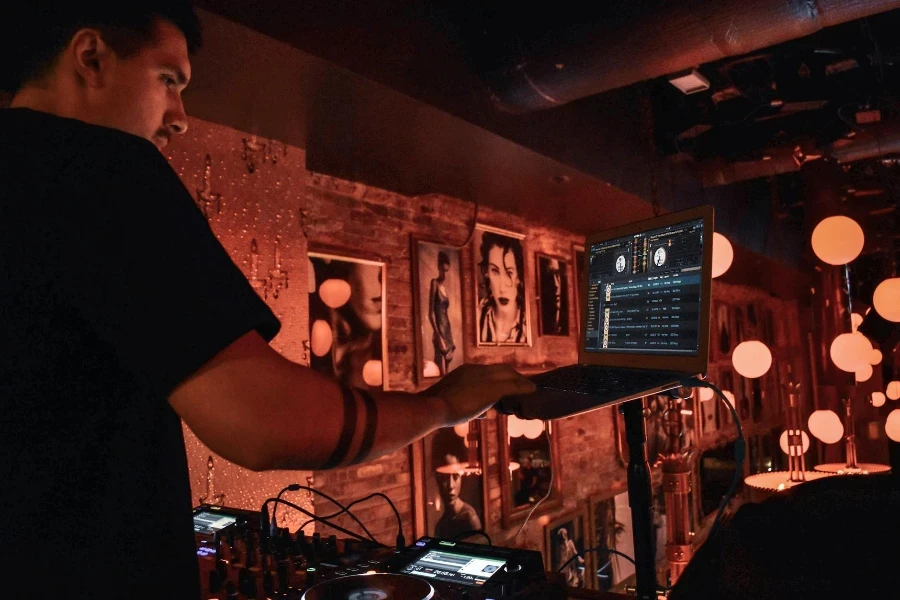 Male DJ in black tee shirt standing behind equipment and selecting songs on laptop