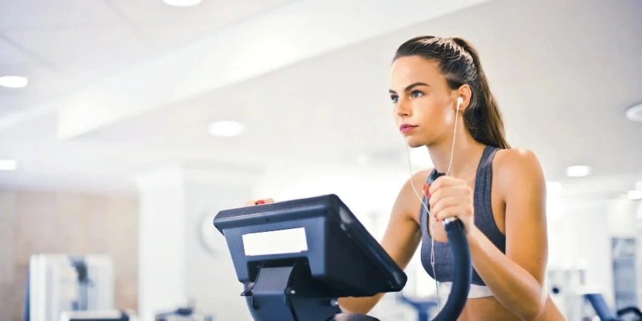 Serious fit woman in earphones and activewear listening to music and running on treadmill in light contemporary sports center