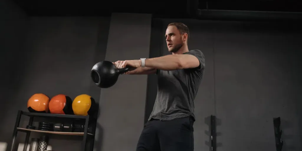 Strong muscular athlete doing kettlebell swings at gym, outfit shot on grey gym background with equipment
