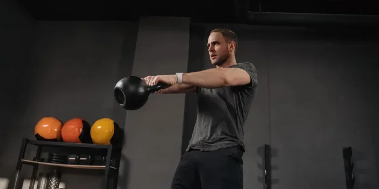 Strong muscular athlete doing kettlebell swings at gym, outfit shot on grey gym background with equipment