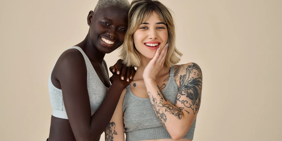 Two happy diverse fit women, Black and European young sporty gen z girls friends multiethnic models wearing sportswear tops standing smiling looking at camera on beige background, portrait