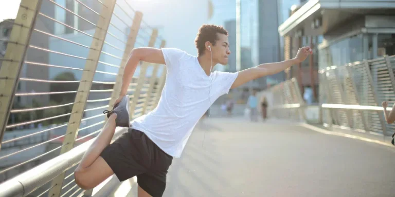Young African American athlete in earbuds listening to music and stretching legs while training alone on street
