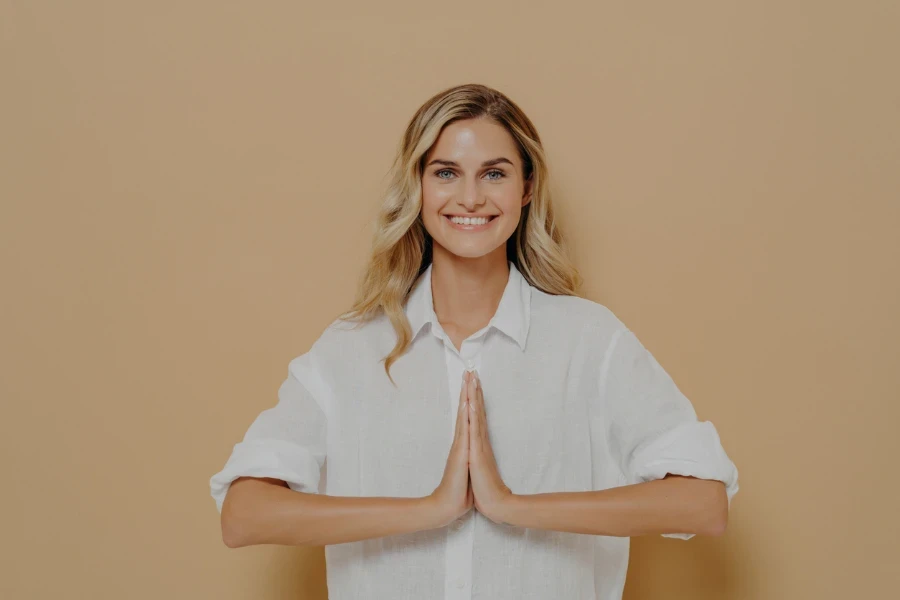 Happy cute blond feamle in white shirt pressing palms together in pray, namaste or saying please gesture, smiling gratefuly and thanking for help, isolated over beige studio background