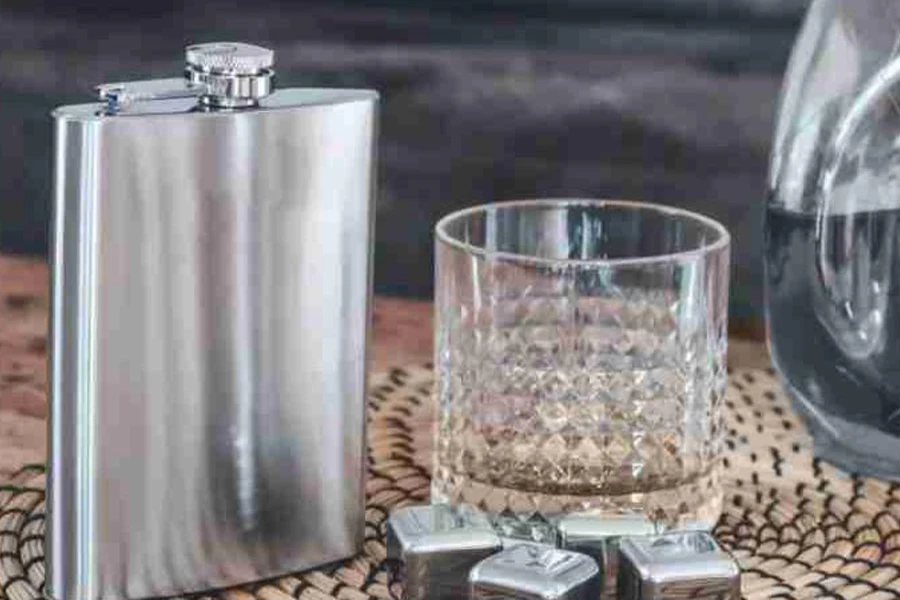 A stainless steel hip flask next to a glass