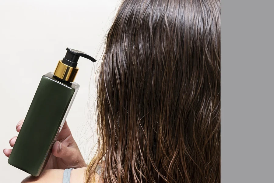 A woman holds a bottle of shampoo or hair balm on her shoulder next to her wet hair