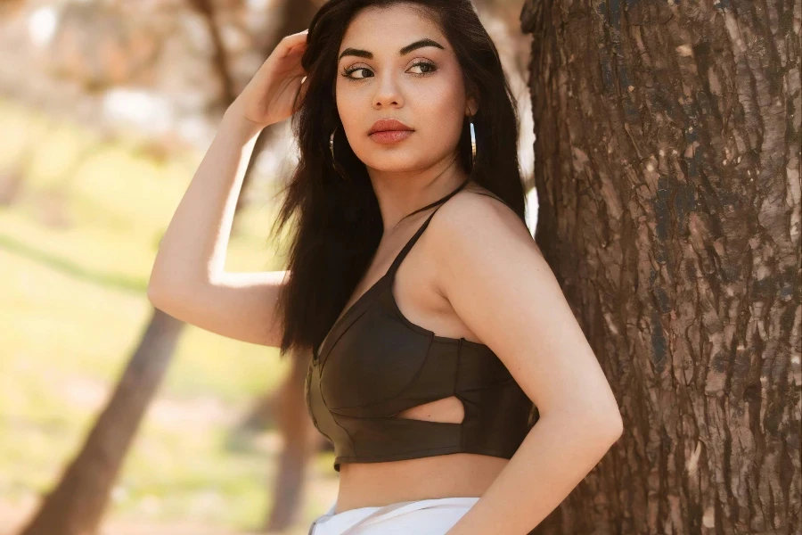 A woman in a crop top and pants leaning against a tree