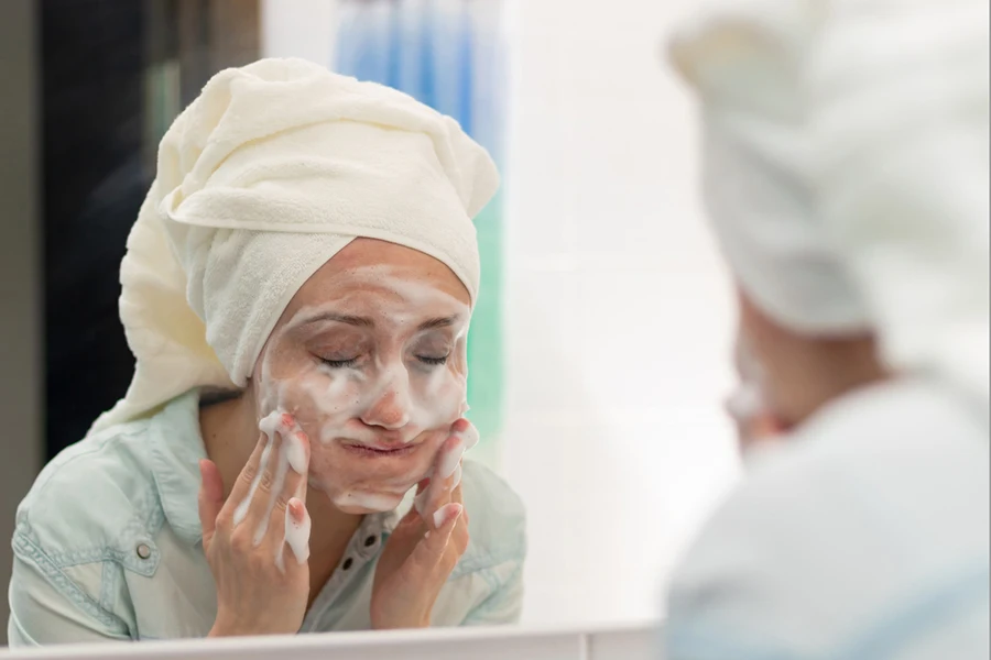 A young woman with a white towel on her head in the bathroom washes off a facial mask