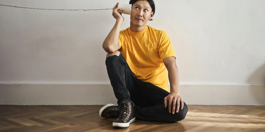 Asian guy in casual clothes using paper cup with thread as telephone while sitting on floor against white wall by Andrea Piacquadio