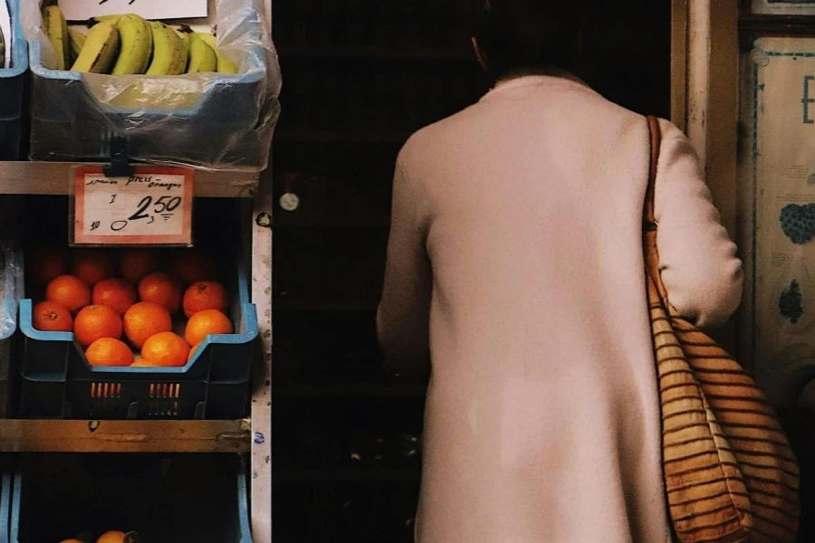 Back View Shot of a Woman Wearing Trench Coat Carrying Bag while Standing Beside Fruit Stand