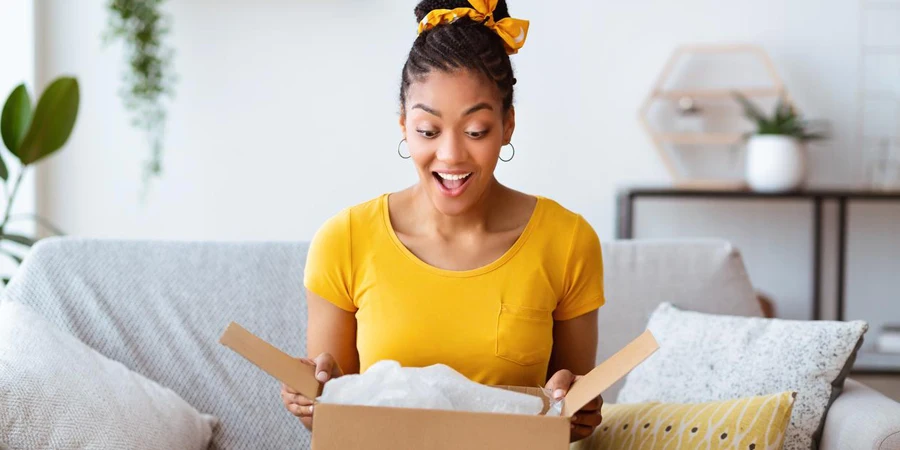 Buying Via Internet. Excited afro girl sitting on sofa unboxing cardboard delivery package, copy space