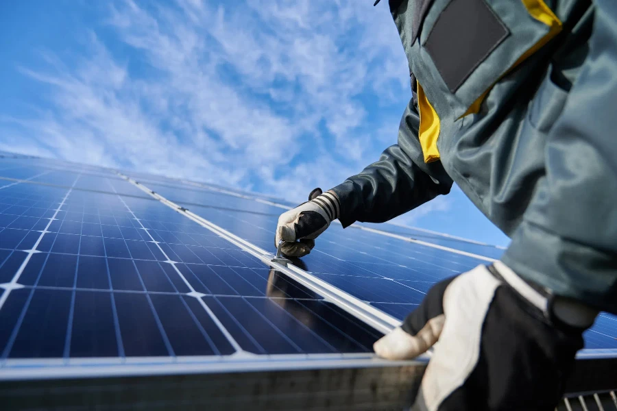 Close up of man technician in work gloves installing stand-alone photovoltaic solar panel system under beautiful blue sky with clouds