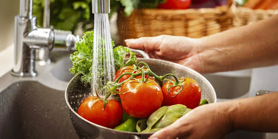 Close up of woman's hands holding a colander full of fresh vegetables under running water in kitchen sink