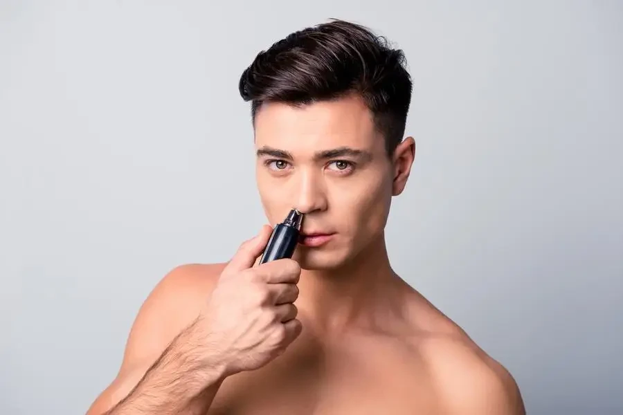 Close up portrait of handsome confident concentrated shaven guy with modern hairdo using a pocket machine for removing unnecessary hair from nose