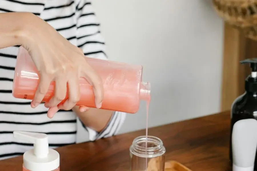 Femme asiatique en tenue décontractée versant du savon couleur pêche d'un distributeur de savon ouvert dans une bouteille en plastique sur un plateau tout en étant assise à table par Sarah Chai