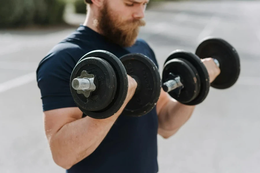 Crop sporty male making effort doing exercise with dumbbells while working out on blurred background of sports ground 