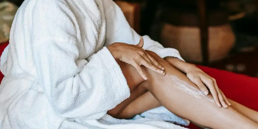 Crop unrecognizable young ethnic female with eye patch applying moisturizing cream on leg while sitting on soft couch by Sora Shimazaki