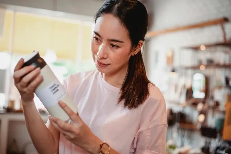 Crop young attentive ethnic shopper reading information on bottle of cosmetic product in store by Sam Lion