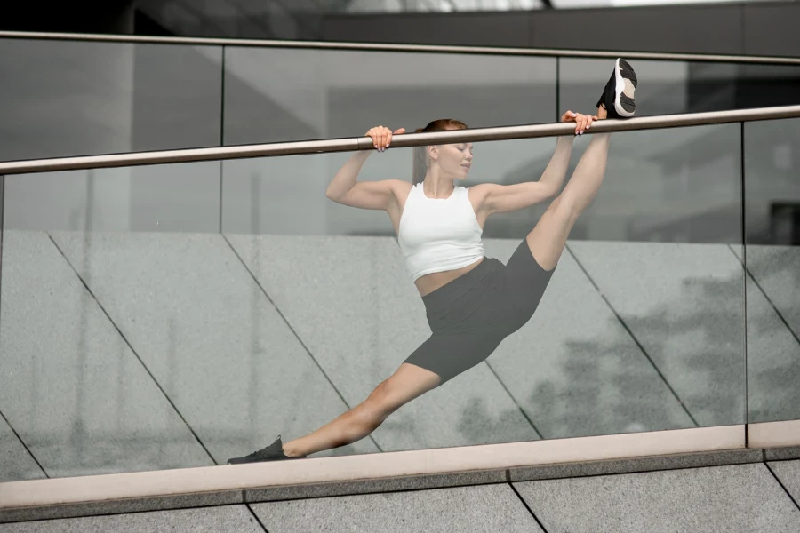 Cute young woman in fitness clothes doing splits stretching near transparent glass railing near building