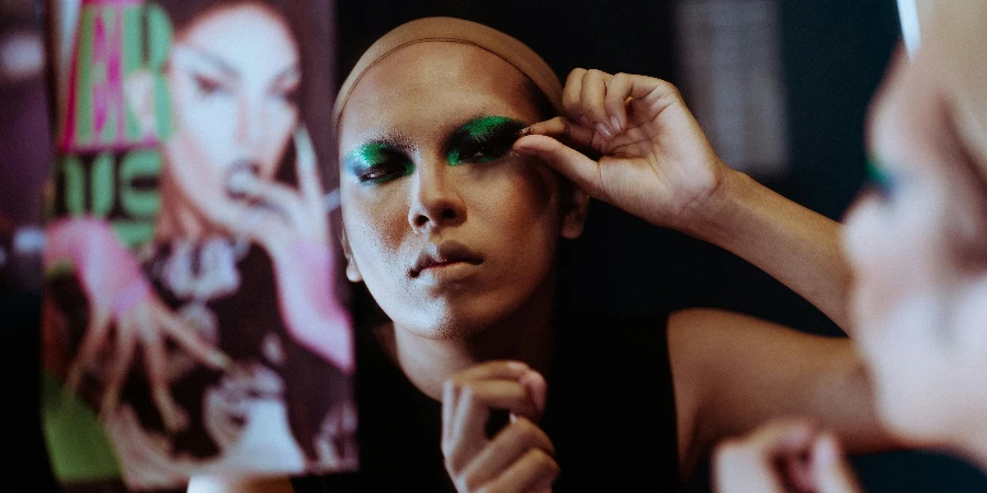 Ethnic female artist in wig cap doing bright makeup with false lashes while looking at mirror reflection