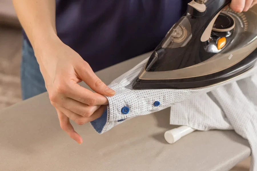 Female hands iron the shirt sleeve on a stand on an ironing board. Ironed clothes lie on the table in the background