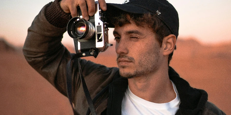 Focused young male traveler in stylish outfit and cap taking photo on vintage camera during trip in desert valley during sunset