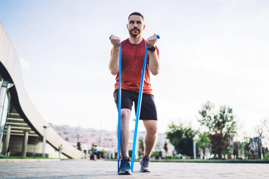 From below of serious bearded male athlete in sportswear having workout while exercising with resistance band outside in summer day