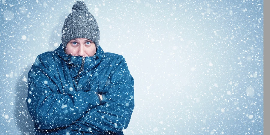 Frozen man in a blue jacket and hat stands against the wall, snow is falling around