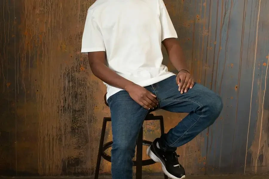 Full body of self assured young African American male model with dreadlocks in casual white t shirt and jeans sitting on chair and looking at camera against wooden wall by Monstera Production