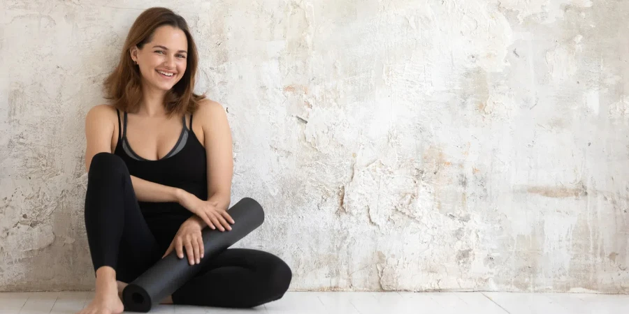 Happy woman wearing sportswear holding black mat, sitting on wooden floor in yoga studio