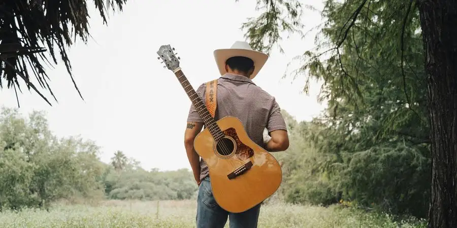 Man in Hat Standing with Guitar on Meadow in Forest by Eli Villarreal