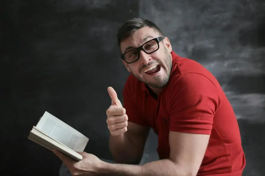 Man in Red Crew Neck T-shirt Wearing Black Framed Eyeglasses Holding White Book by Andrea Piacquadio