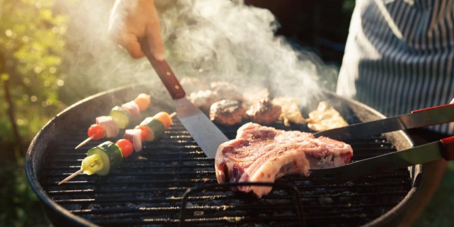 Man with drink cooking meat and vegetables on barbecue grill outdoors, closeup