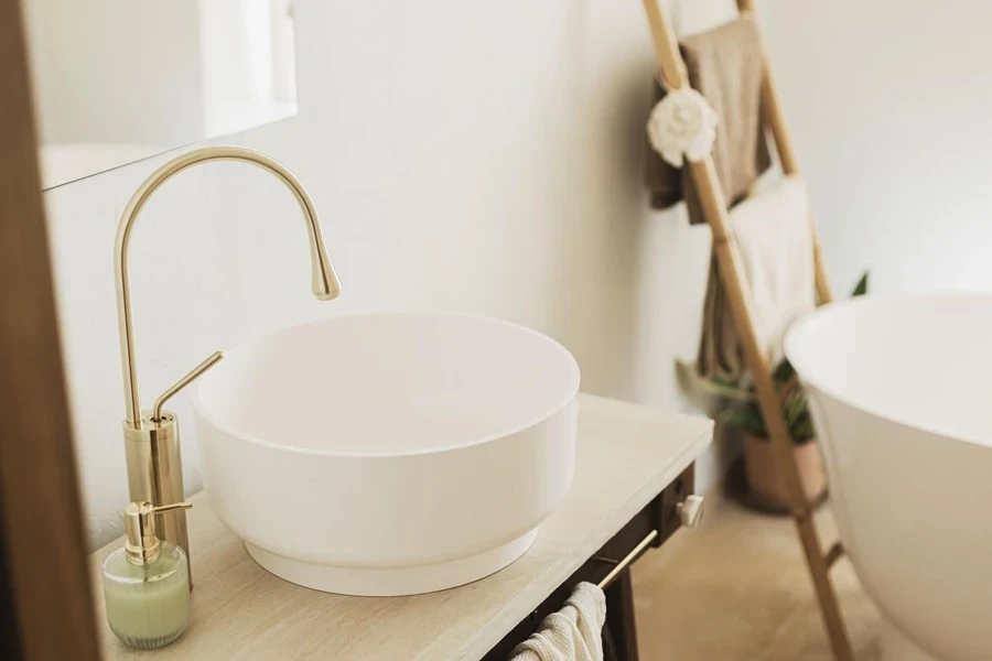 Modern ceramic sink with golden faucet on rustic stand with soap and towel on background of bathtub, modern bathroom interior. 