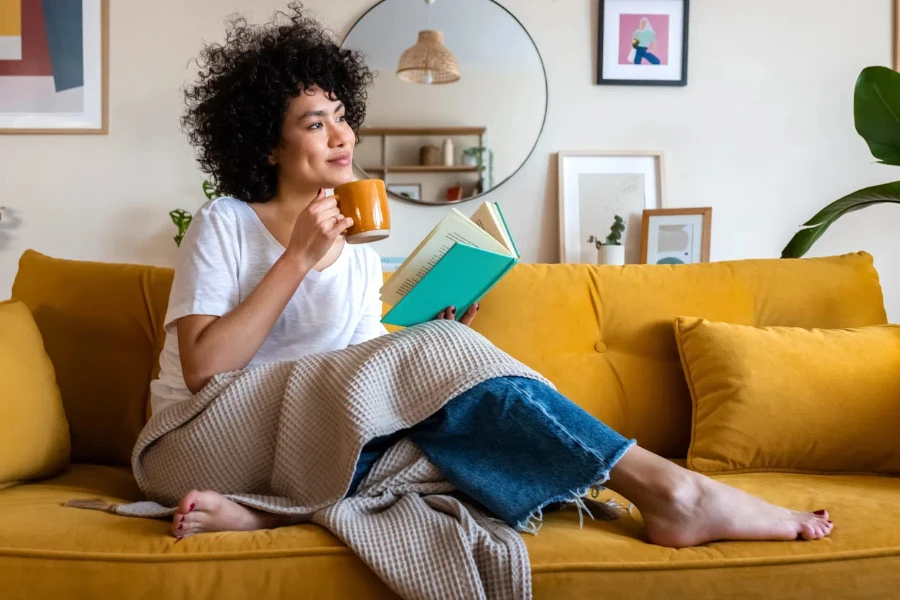 Pensive relaxed African american woman reading a book at home, drinking coffee sitting on the couch. Copy space. Lifestyle concept