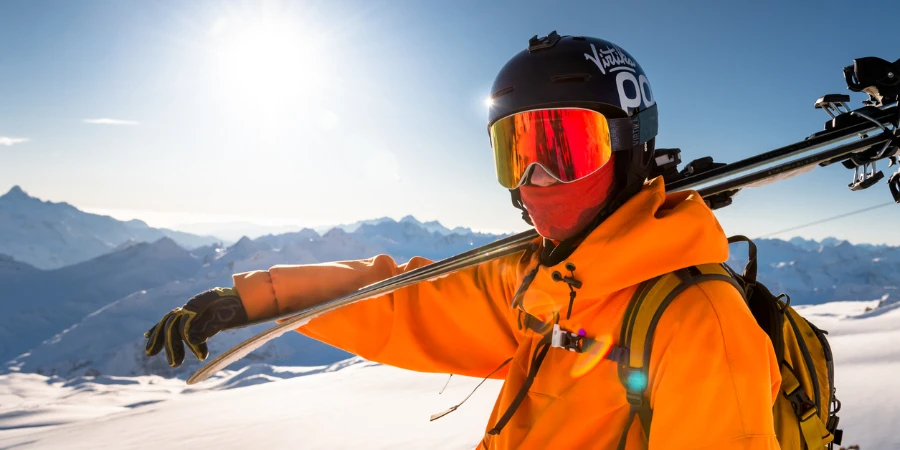 Portrait of a confident skier in an orange jacket standing on the top of a mountain and holding skis in his hands