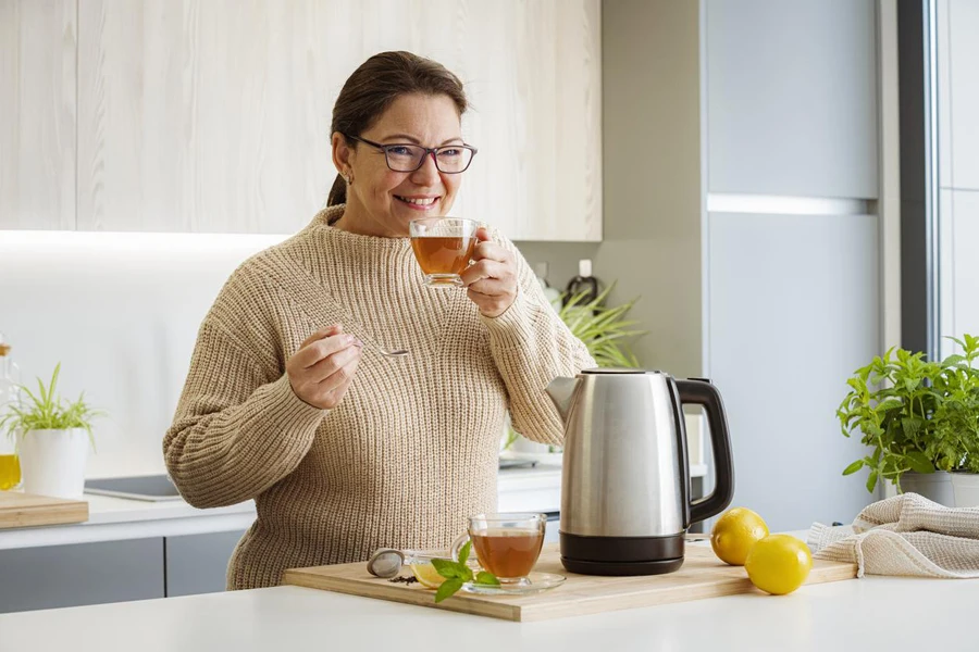 Portrait of a woman drinking hot tea in the kitchen. An electric kettle, mint plant in a pot and limes complete the composition.