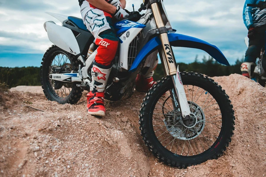 Racer in sportswear sitting on motorbike on sandy pile against cloudy sky with forest in distance in daylight