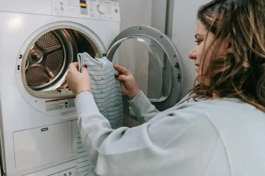 Side view of crop female in casual outfit looking at baby bodysuit before washing clothes in laundry room by Sarah Chai