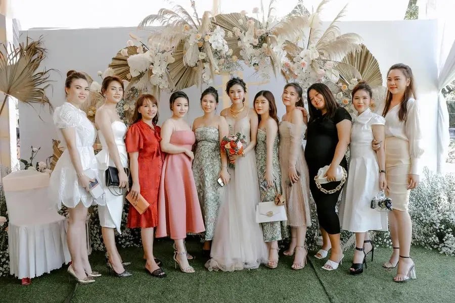 Smiling Asian woman in elegant dress standing near friends and looking at camera after wedding ceremony by Trung Nguyen