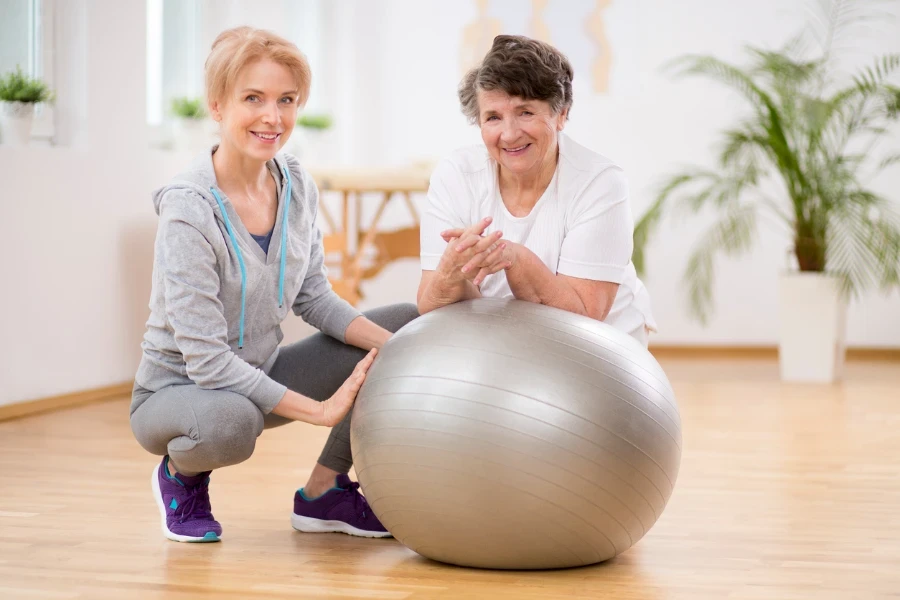 Smiling physiotherapist with elderly woman laying on exercising ball during physical therapy