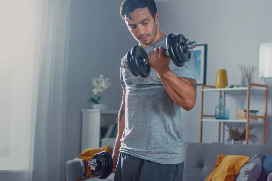 Strong Athletic Fit Man in T-shirt and Shorts is Doing Calf Raise Exercises with Dumbbells at Home in His Spacious and Bright Apartment with Minimalistic Interior. 